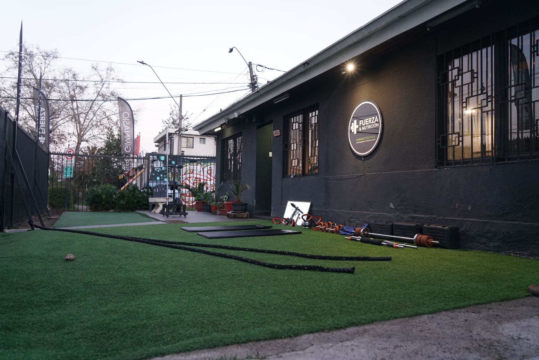Fachada de Fuerza & Nutrición desde la vereda: patio de pasto sintético con barras y cuerdas, y el letrero del gimnasio